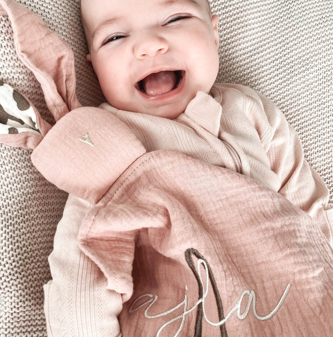 Smiling baby holding a pink personalised bunny comforter with embroidered name. Ideal for soothing and cuddling.