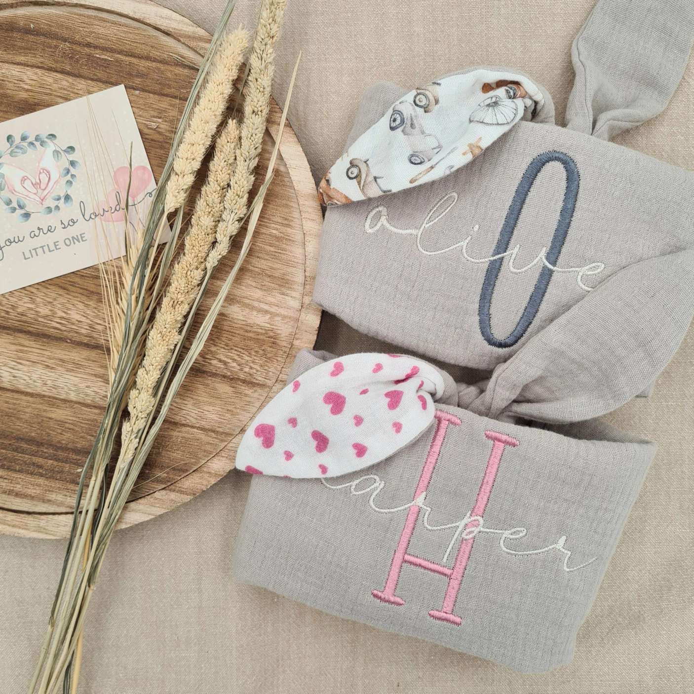 Grey baby comforters with pink and grey patterned bunny ears, embroidered with names 'Olive' and 'Harper'. Soft, comforting, and personalised.